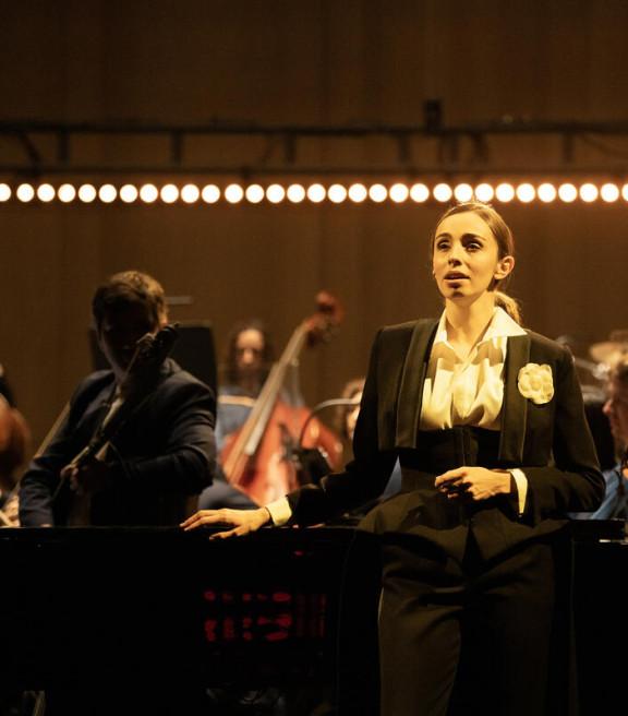 Lea Desandre est adossée au piano, en tenue de queue de pie. Derrière elle, on aperçoit une partie de l'orchestre